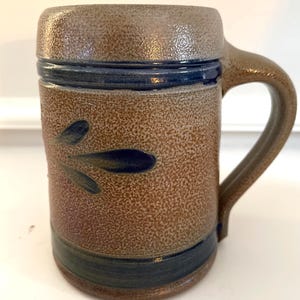 May include: A brown stoneware mug with a textured surface and a handle. The mug features a dark blue band around the top and bottom, with a decorative leaf design in dark blue. The mug is approximately 5 inches tall.
