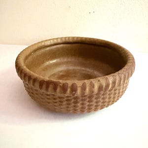 May include: A round, medium-brown ceramic bowl with a textured exterior featuring a repeating dimpled pattern. The rim of the bowl is thick and rounded, displaying a series of vertical indentations that create a decorative, scalloped edge.
