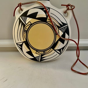 May include: A white ceramic canteen with a black and yellow geometric design and a brown leather strap.