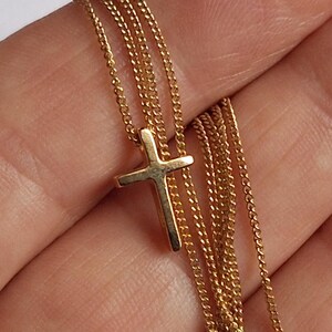 Puede incluir: Un collar con colgante de cruz de color dorado. La cruz es peque&ntilde;a y cuelga de una delicada cadena. El collar se muestra sobre un fondo neutro.