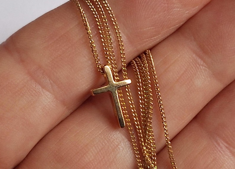 Puede incluir: Un collar con colgante de cruz de color dorado. La cruz es peque&ntilde;a y cuelga de una delicada cadena. El collar se muestra sobre un fondo neutro.