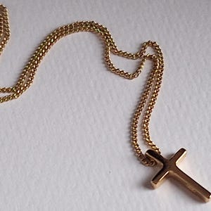 Puede incluir: Un collar dorado con un colgante de cruz. La cadena tiene un dise&ntilde;o delicado y enlazado. La cruz tiene una forma simple y cl&aacute;sica. El collar se muestra sobre una superficie blanca.