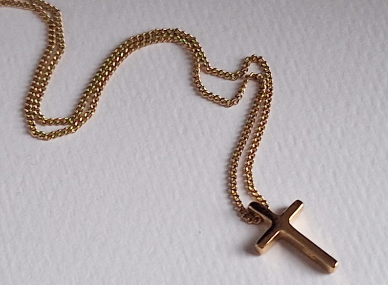 Puede incluir: Un collar dorado con un colgante de cruz. La cadena tiene un dise&ntilde;o delicado y enlazado. La cruz tiene una forma simple y cl&aacute;sica. El collar se muestra sobre una superficie blanca.