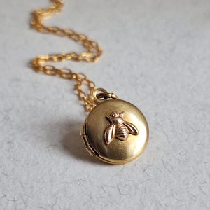 May include: A gold locket necklace featuring a bee design. The locket is round and has a hinged lid. The bee is crafted from gold and has a raised design.