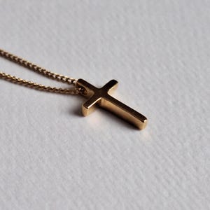 Puede incluir: Un collar con colgante de cruz de color dorado. La cruz tiene un dise&ntilde;o simple y s&oacute;lido con bordes redondeados. La cadena es delicada y del mismo color dorado. El collar se muestra sobre una superficie blanca.