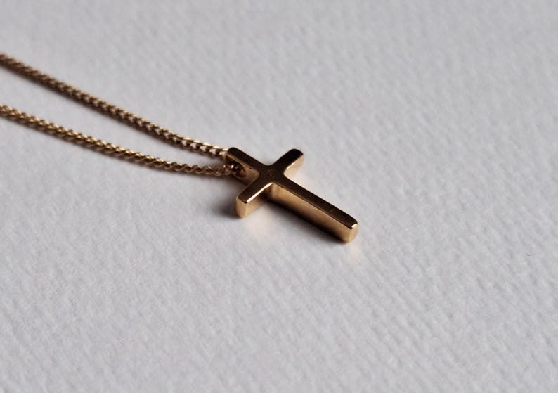 Puede incluir: Un collar con colgante de cruz de color dorado. La cruz tiene un dise&ntilde;o simple y s&oacute;lido con bordes redondeados. La cadena es delicada y del mismo color dorado. El collar se muestra sobre una superficie blanca.