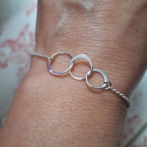 Könnte beinhalten: Ein silbernes Armband mit drei miteinander verbundenen, offenen Kreisen. Das Armband ist an einer filigranen Kette befestigt und wird an einem Handgelenk gezeigt. Die Kreise sind glatt und glänzend.