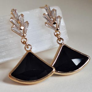 May include: Gold-toned earrings with a geometric design. Each earring features a black, fan-shaped pendant framed in gold, suspended from a cluster of clear, baguette-cut crystals. The earrings are designed for pierced ears.