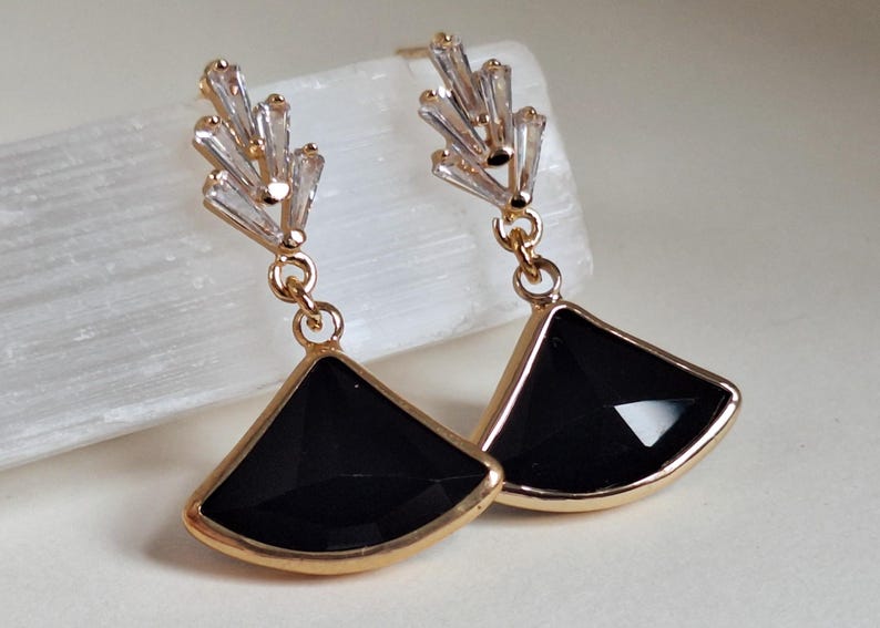 May include: Gold-toned earrings with a geometric design. Each earring features a black, fan-shaped pendant framed in gold, suspended from a cluster of clear, baguette-cut crystals. The earrings are designed for pierced ears.
