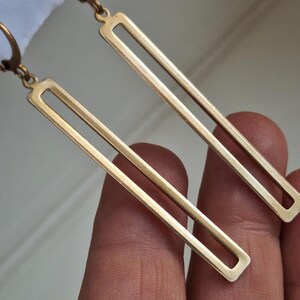 Puede incluir: Un par de pendientes rectangulares dorados. Cada pendiente tiene un dise&ntilde;o de rect&aacute;ngulo largo y abierto. Los pendientes cuelgan de un gancho. El fondo es una vista borrosa de una mano que sostiene los pendientes.
