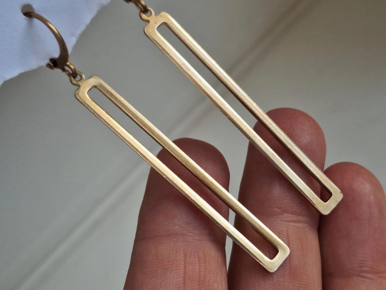 Puede incluir: Un par de pendientes rectangulares dorados. Cada pendiente tiene un dise&ntilde;o de rect&aacute;ngulo largo y abierto. Los pendientes cuelgan de un gancho. El fondo es una vista borrosa de una mano que sostiene los pendientes.
