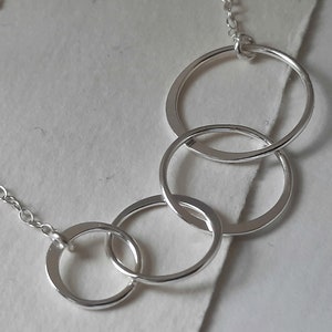 Peut inclure: Un collier en argent composé de quatre cercles ouverts et interconnectés. Les cercles sont liés, créant un design moderne et minimaliste. Le collier est attaché à une chaîne délicate.