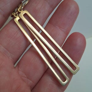 Puede incluir: Un par de pendientes colgantes rectangulares dorados. Cada pendiente presenta tres rect&aacute;ngulos paralelos y abiertos. Los pendientes est&aacute;n suspendidos de un peque&ntilde;o gancho dorado. Los pendientes se sostienen en una mano.