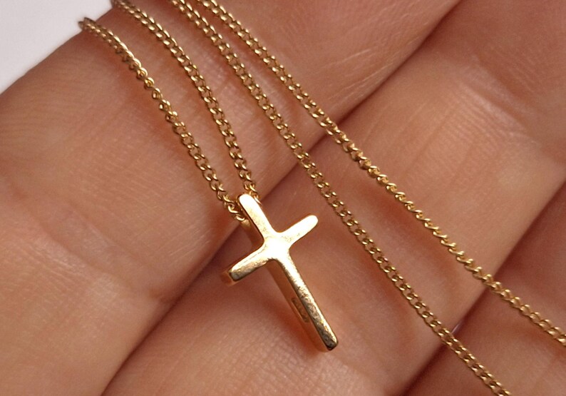 Puede incluir: Un collar dorado con un peque&ntilde;o colgante de cruz. La cruz tiene un dise&ntilde;o sencillo y cl&aacute;sico, suspendido de una delicada cadena. El collar se muestra sobre un fondo neutro, resaltando los detalles de la joya.