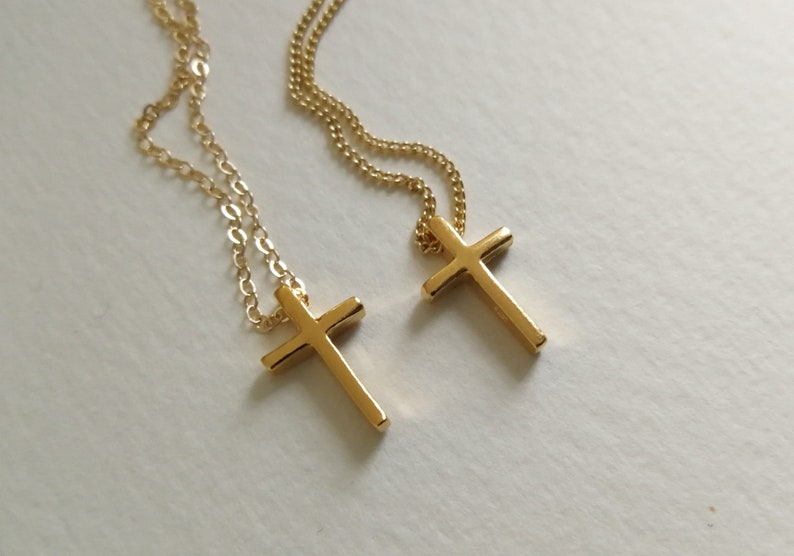 Puede incluir: Dos collares con cruces doradas. Cada collar presenta una cadena delicada y un colgante de cruz simple y pulido. Las cruces est&aacute;n centradas y tienen una superficie lisa y reflectante. El fondo es de color blanco roto.