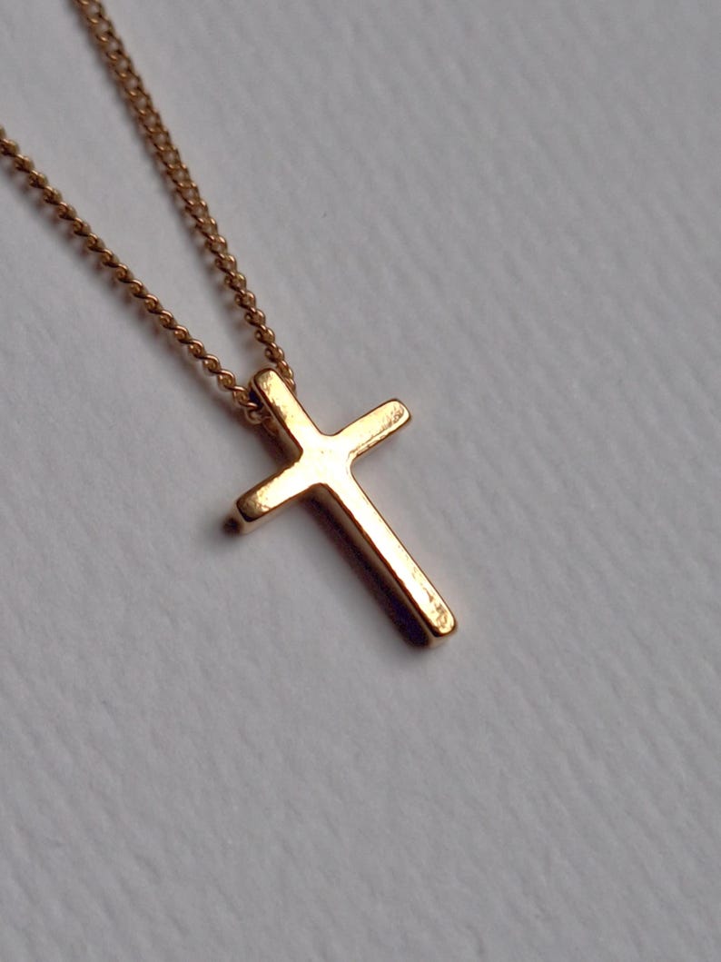 Puede incluir: Un collar con colgante de cruz de color dorado. La cruz tiene un dise&ntilde;o simple y cl&aacute;sico con bordes redondeados. La cadena es delicada y tambi&eacute;n dorada. El collar se muestra sobre un fondo suave y neutro.