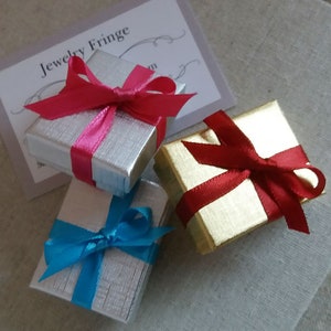 May include: Three small gift boxes, two silver and one gold, are tied with colorful ribbons. The silver boxes have pink and blue ribbons, while the gold box has a red ribbon. A card with the text "Jewelry Fringe" is visible.
