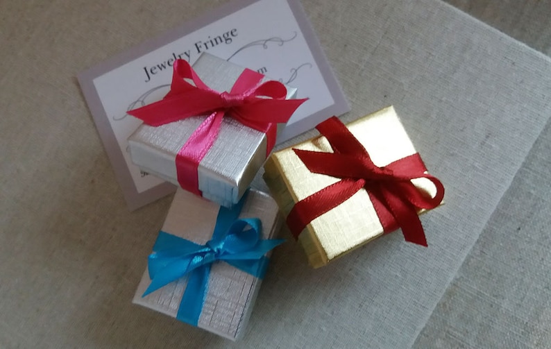 May include: Three small gift boxes, two silver and one gold, are tied with colorful ribbons. The silver boxes have pink and blue ribbons, while the gold box has a red ribbon. A card with the text "Jewelry Fringe" is visible.