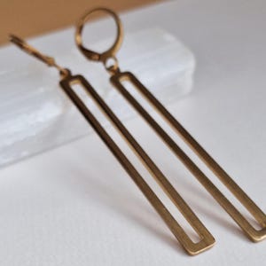 Puede incluir: Un par de pendientes largos y rectangulares de lat&oacute;n con un dise&ntilde;o geom&eacute;trico simple. Los pendientes tienen un cierre de palanca y un acabado pulido. Los pendientes se exhiben sobre una superficie blanca.