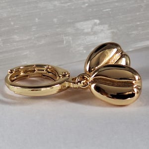 Könnte beinhalten: Goldfarbene Kaffeebohnen-Ohrringe. Die Ohrringe verfügen über einen Klappbügelverschluss und drei detaillierte Kaffeebohnen-Anhänger. Der Schmuck ist vor einem weichen, neutralen Hintergrund platziert, der das polierte Finish des Metalls hervorhebt.