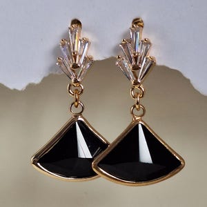 May include: A pair of gold-toned earrings with a fan-shaped black stone pendant. The earrings have a cluster of clear, rectangular crystals at the top. The earrings are hanging against a neutral background.