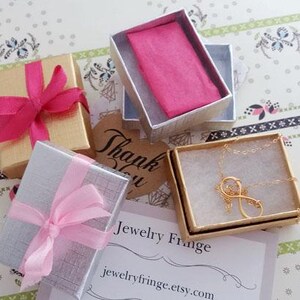 Puede incluir: Vista a&eacute;rea de joyeros, algunos con lazos rosas. Una caja contiene un collar dorado con forma de s&iacute;mbolo de infinito. Una peque&ntilde;a etiqueta dice "Gracias". Las cajas est&aacute;n sobre una superficie con estampado floral.