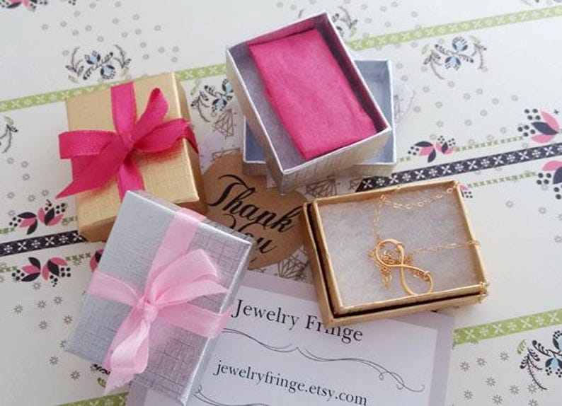 Puede incluir: Vista a&eacute;rea de joyeros, algunos con lazos rosas. Una caja contiene un collar dorado con forma de s&iacute;mbolo de infinito. Una peque&ntilde;a etiqueta dice "Gracias". Las cajas est&aacute;n sobre una superficie con estampado floral.