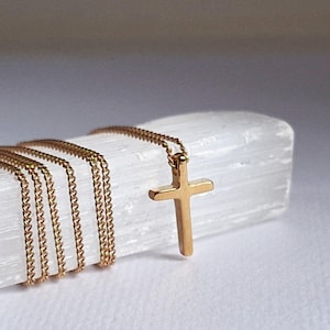 Puede incluir: Un collar con colgante de cruz de color dorado con una cadena delicada. La cruz es simple y elegante, y la cadena est&aacute; envuelta alrededor de un objeto blanco texturizado. El collar se muestra sobre un fondo blanco.