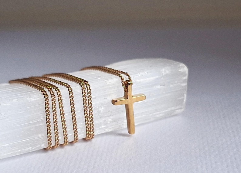 Puede incluir: Un collar con colgante de cruz de color dorado con una cadena delicada. La cruz es simple y elegante, y la cadena est&aacute; envuelta alrededor de un objeto blanco texturizado. El collar se muestra sobre un fondo blanco.