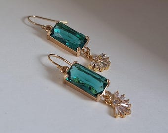 May include: A pair of gold-colored earrings with emerald-green rectangular gems. Each earring has a small, gold-colored floral accent with clear, rectangular crystals. The earrings are displayed on a white surface.