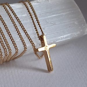 Puede incluir: Un collar con colgante de cruz dorada. La cruz, de dise&ntilde;o cl&aacute;sico, cuelga de una delicada cadena. La joya se exhibe sobre una superficie blanca y texturizada, resaltando el brillo del oro.