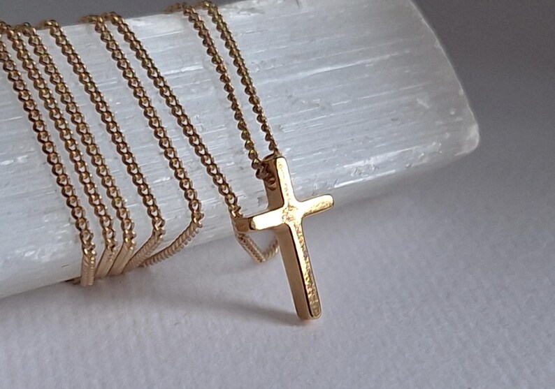 Puede incluir: Un collar con colgante de cruz dorada. La cruz, de dise&ntilde;o cl&aacute;sico, cuelga de una delicada cadena. La joya se exhibe sobre una superficie blanca y texturizada, resaltando el brillo del oro.