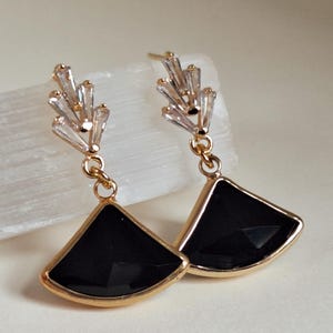May include: A pair of gold-toned earrings with a fan-shaped black stone pendant. The earrings have a cluster of clear, rectangular crystals at the top. The earrings are hanging.