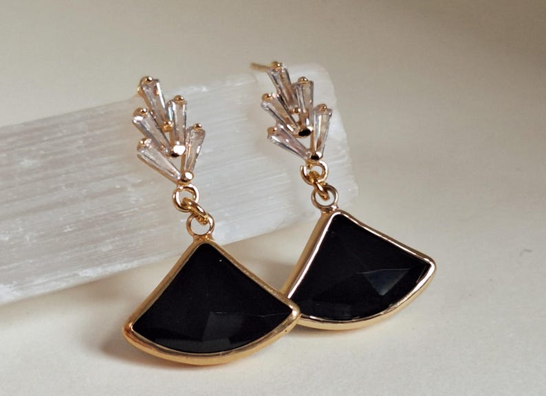 May include: A pair of gold-toned earrings with a fan-shaped black stone pendant. The earrings have a cluster of clear, rectangular crystals at the top. The earrings are hanging.