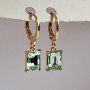 May include: Gold-coloured hoop earrings featuring rectangular, pale green gemstones. The earrings are presented against a neutral backdrop, highlighting their design and colour.