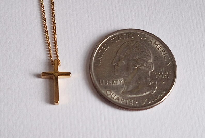 Puede incluir: Un collar con colgante de cruz de color dorado y una cadena delicada. La cruz es peque&ntilde;a y de dise&ntilde;o sencillo. Una moneda de veinticinco centavos estadounidense est&aacute; junto al collar para comparar el tama&ntilde;o.