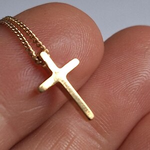 Puede incluir: Un peque&ntilde;o colgante de cruz de color dorado en una delicada cadena. La cruz tiene un dise&ntilde;o simple y cl&aacute;sico, y la cadena parece ser de estilo cuerda trenzada. La joya se sostiene entre los dedos.