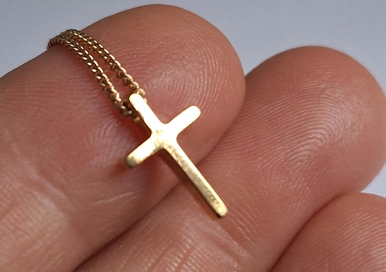 Puede incluir: Un peque&ntilde;o colgante de cruz de color dorado en una delicada cadena. La cruz tiene un dise&ntilde;o simple y cl&aacute;sico, y la cadena parece ser de estilo cuerda trenzada. La joya se sostiene entre los dedos.