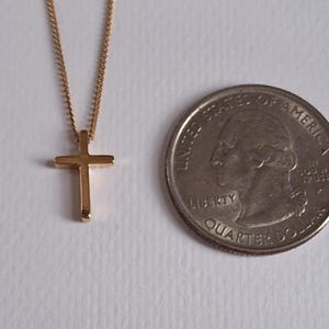 Puede incluir: Un collar con colgante de cruz de color dorado. La cruz es peque&ntilde;a y sencilla, suspendida de una delicada cadena. Una moneda de veinticinco centavos estadounidense est&aacute; al lado del collar para comparar el tama&ntilde;o.