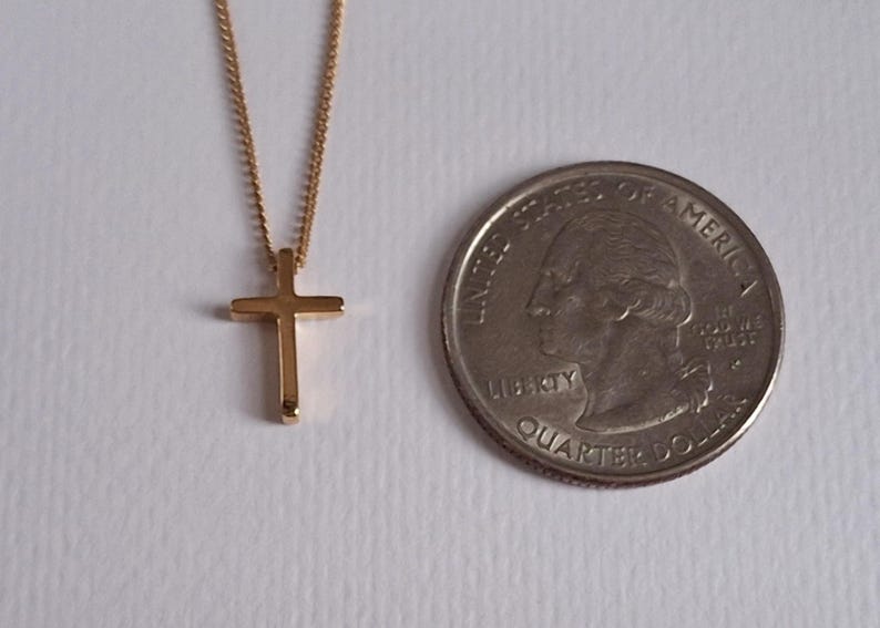 Puede incluir: Un collar con colgante de cruz de color dorado. La cruz es peque&ntilde;a y sencilla, suspendida de una delicada cadena. Una moneda de veinticinco centavos estadounidense est&aacute; al lado del collar para comparar el tama&ntilde;o.