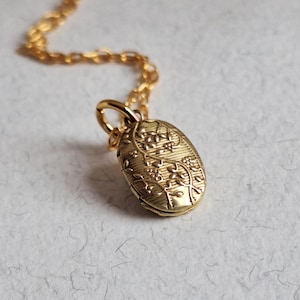 Può includere: Una collana con pendente ovale dorato con un motivo floreale. Il pendente è sospeso a una delicata catena d'oro. La collana è esposta su una superficie di colore chiaro.