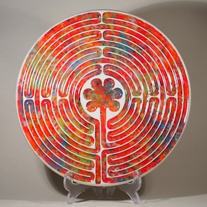 Peut inclure: Une assiette décorative ronde avec un motif de labyrinthe coloré. Le bord est blanc et un motif floral central est présent. Le labyrinthe est composé d'anneaux concentriques aux teintes de rouge, orange, jaune et bleu.