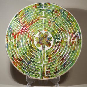 May include: A circular ceramic plate with a labyrinth pattern. The plate has a white base with a green, splattered paint design. A flower design is in the centre. The plate is displayed on a clear plastic stand.