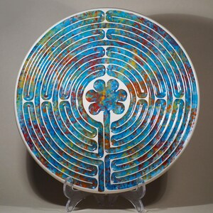 May include: A circular decorative plate with a labyrinth pattern. The plate has a white base with a blue, red, and yellow design. A flower shape is at the centre. The plate is displayed on a clear stand.