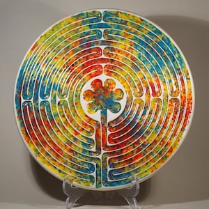 Peut inclure: Une assiette décorative ronde et colorée avec un motif de labyrinthe. L'assiette a un bord blanc et une forme centrale en forme de fleur. Le labyrinthe est composé de chemins concentriques aux teintes vives de rouge, jaune et bleu.