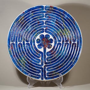 May include: A circular ceramic plate featuring a blue labyrinth pattern. The design consists of concentric paths outlined in white. The centre has a flower motif with red, yellow, and blue details. The plate is presented on a clear stand.