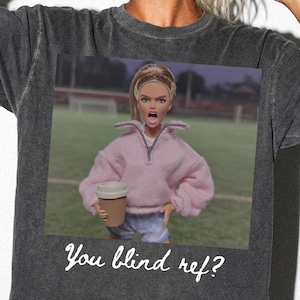 May include: A charcoal gray short-sleeve t-shirt featuring a graphic print of a doll with blonde hair wearing a pink quarter-zip sweatshirt and holding a coffee cup on a soccer field. The text "You blind ref?" is printed in white below the graphic.