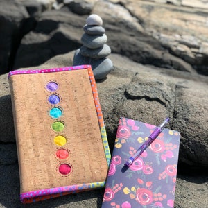 May include: A cork notebook cover with a rainbow of colors in the center and a floral patterned notebook with a pen on top. The text on the notebook says "Field Book".
