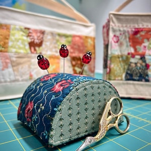 May include: A blue and green fabric pincushion shaped like a half-circle with four red ladybug pins. A gold bird-shaped pair of scissors is lying on a green cutting mat.