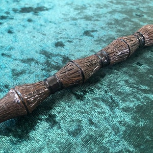 May include: A brown wand with a textured, wood-like appearance. The wand features dark bands and a detailed, carved design. The tip of the wand is slightly pointed. The wand is set against a teal green background.